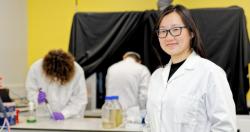Jenny Zhang in white lab coat stands in front of lab bench with Joshua Lawrence and Xiaolong Chen in background.