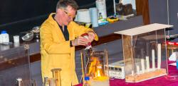 Dr Peter Wothers in lab glasses & yellow lab coat with an array of experiments for the audience