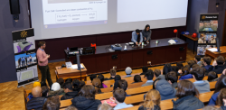 Three presenters perform an experiment at a lecturn in front of an audience of students.