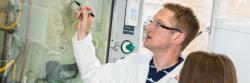 A man in a lab coat writes on a glass fume hood.