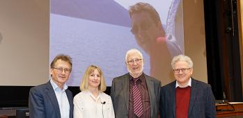 Ben Feringa, Sally Boss, John Hopkins & Jeremy Sanders looking ahead, standing in front of photo of Alex Hopkins on screen