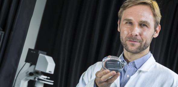 Tuomas Knowles in lab coat holding a microfluidics chip in his hand and looking at camera