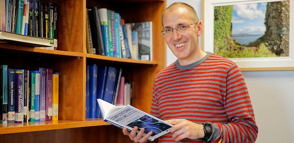 Prof Michaelides standing next to a bookcase holding an open book