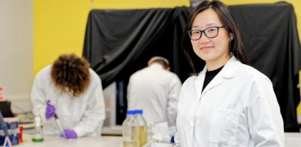 Jenny Zhang in white lab coat stands in front of lab bench with Joshua Lawrence and Xiaolong Chen in background.