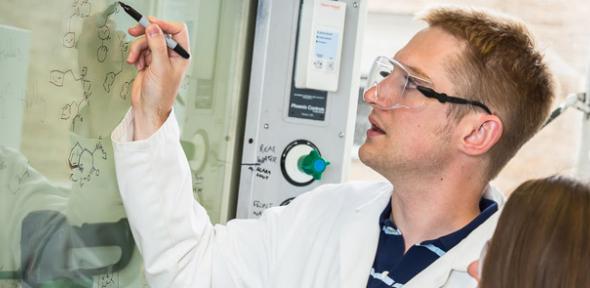 A man in a lab coat writes on a glass fume hood.