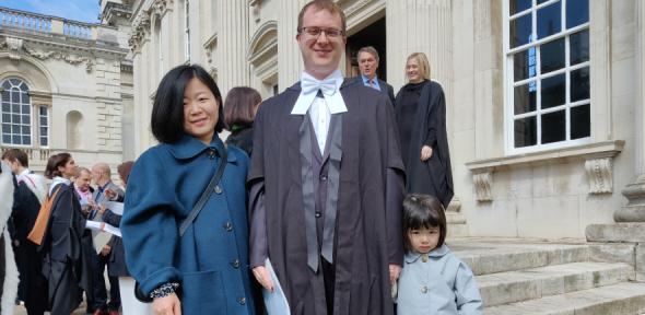 Adam Hall in graduation gown with wife and child smiling