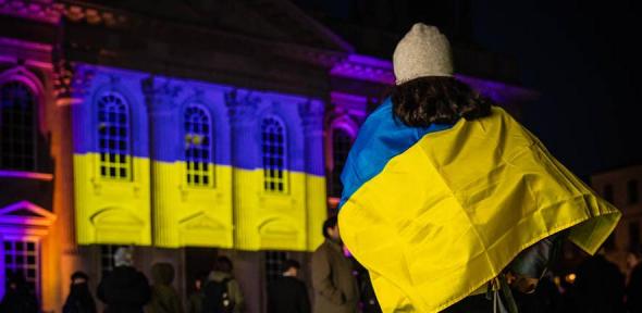 Person wearing Ukrainian flag in front of building with Ukrainian flag projected on the front.