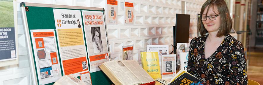 Megan Holman reading a book next to a table in the library with all the exhibition items displayed