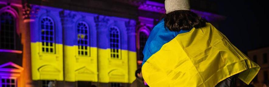 Person wearing Ukrainian flag in front of building with Ukrainian flag projected on the front.